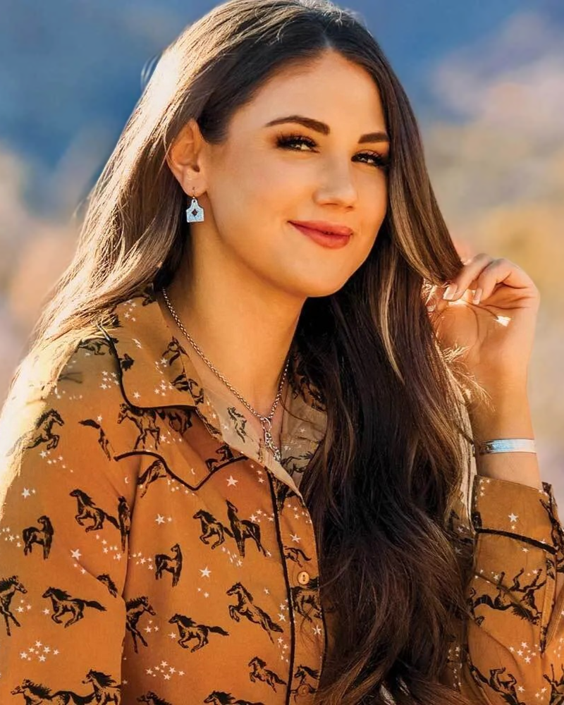 Woman in a patterned shirt and wearing Montana Silversmiths jewelry, standing in a desert landscape with mountains in the background.
